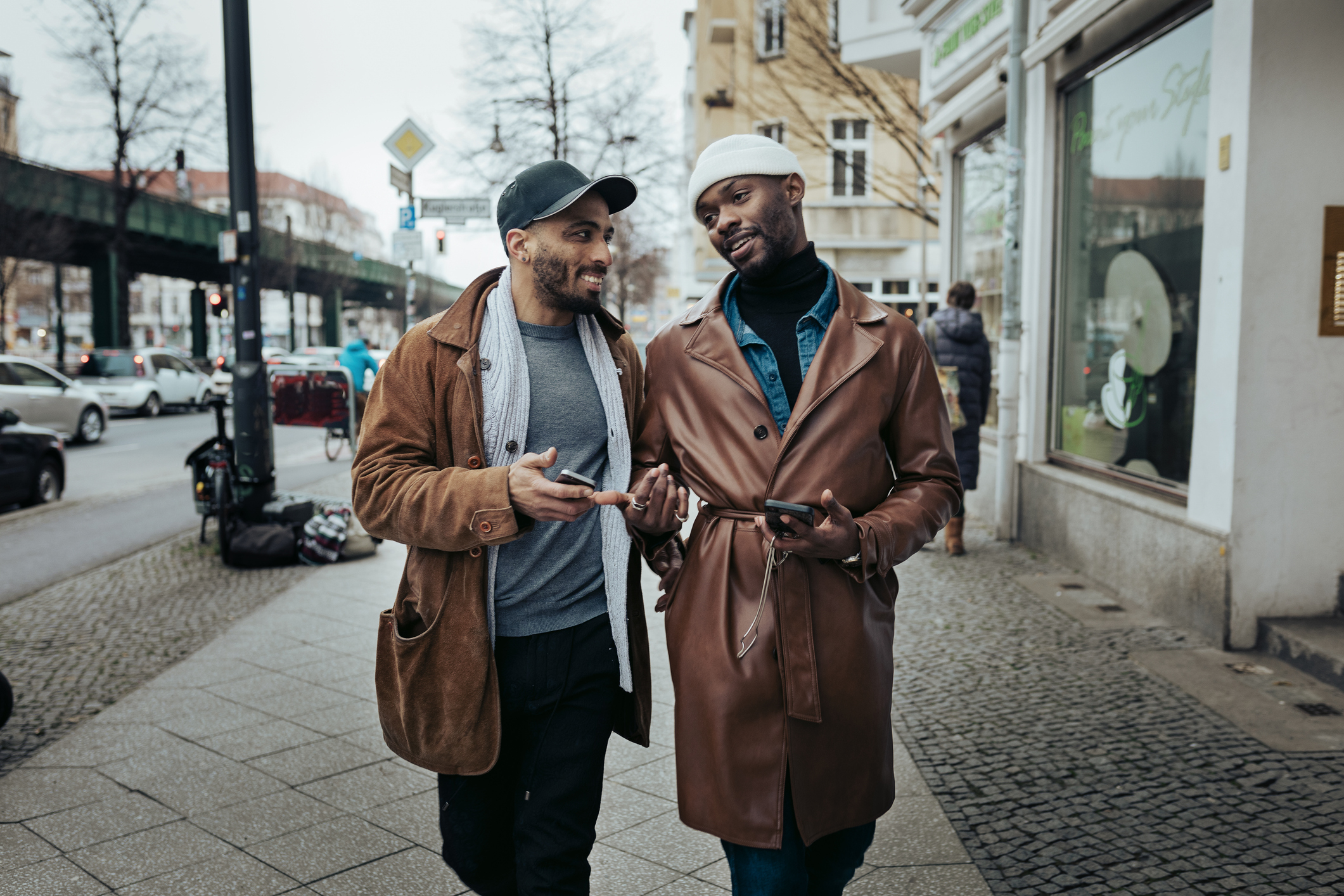 Two men, one in a casual jacket and cap, the other in a stylish leather coat and beanie, walk and converse on a city street with parked bikes and cars