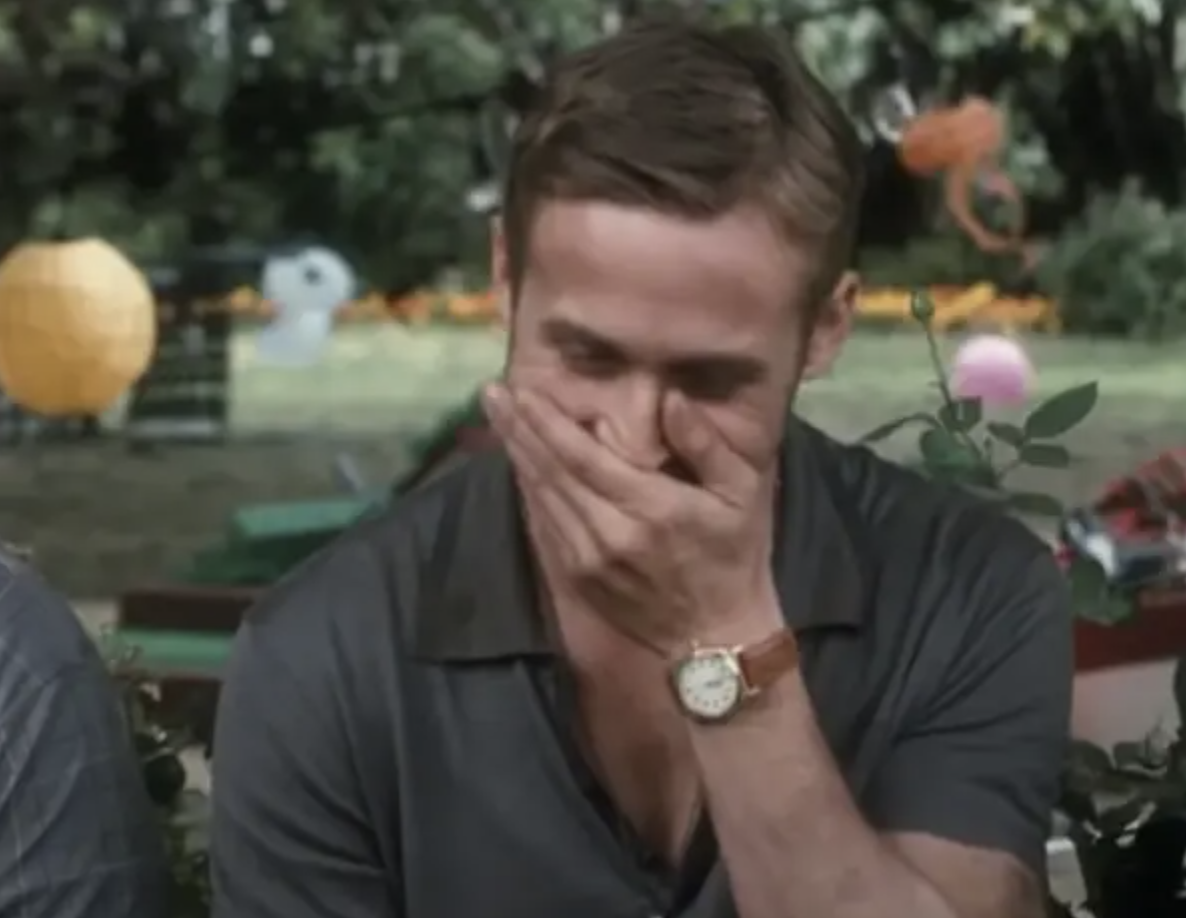 Ryan Gosling sitting outdoors, covering his mouth with his hand, laughing and wearing a casual shirt and a watch