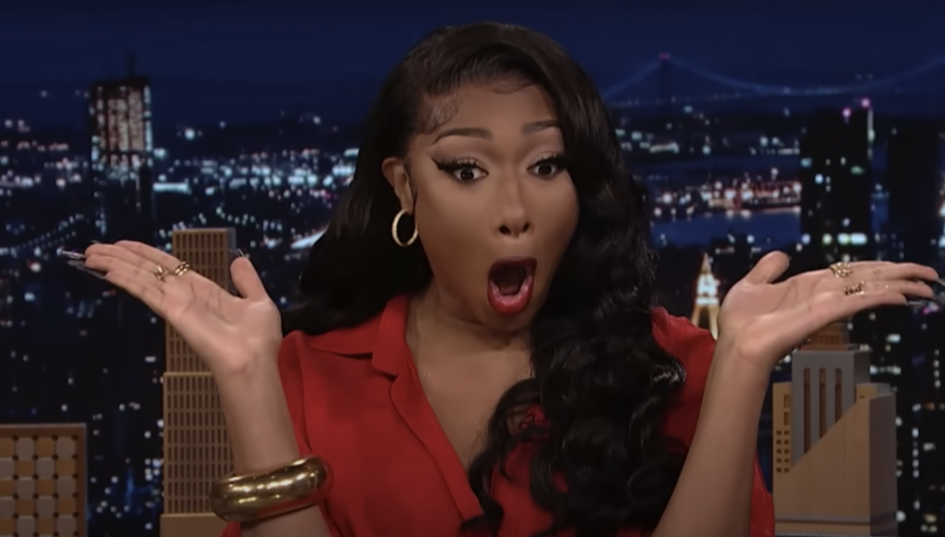 Megan Thee Stallion, wearing a long-sleeve top and large hoop earrings, looks surprised with hands up during a TV interview in front of a cityscape background