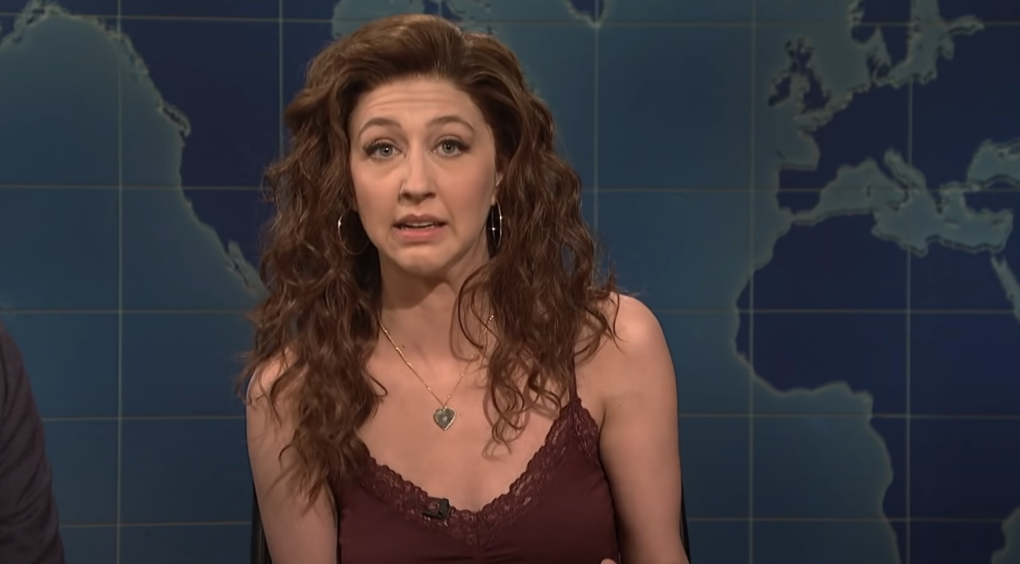 A woman with curly hair wearing a sleeveless top is speaking on a news program. A world map is in the background