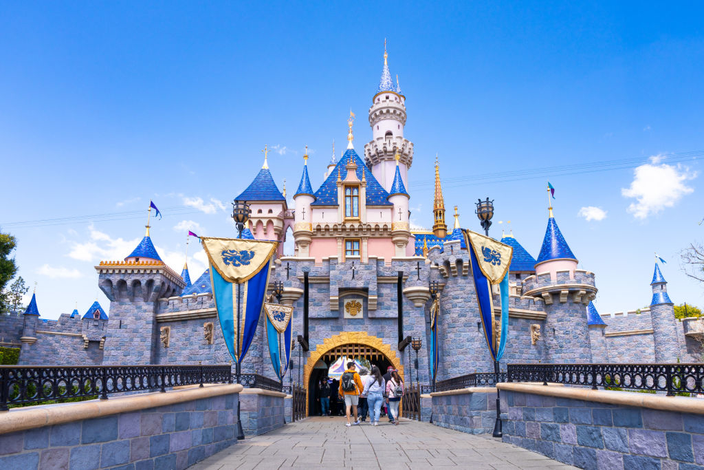 People walking through the entrance of Sleeping Beauty Castle at Disneyland. The castle features spires and flags