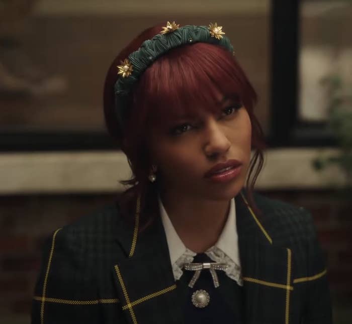 I don't know who this person is. In the image, a person is wearing a plaid blazer with a white lace collar and a jeweled brooch, accessorized with a decorative headband