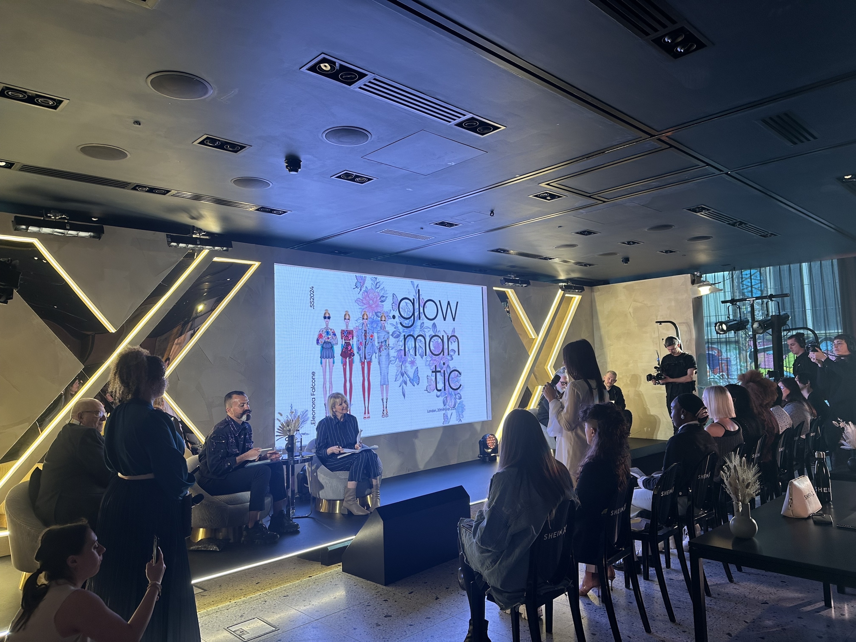 A panel of speakers is presenting &quot;Glow Romantic&quot; at an indoor event. Attendees are seated, observing the presentation
