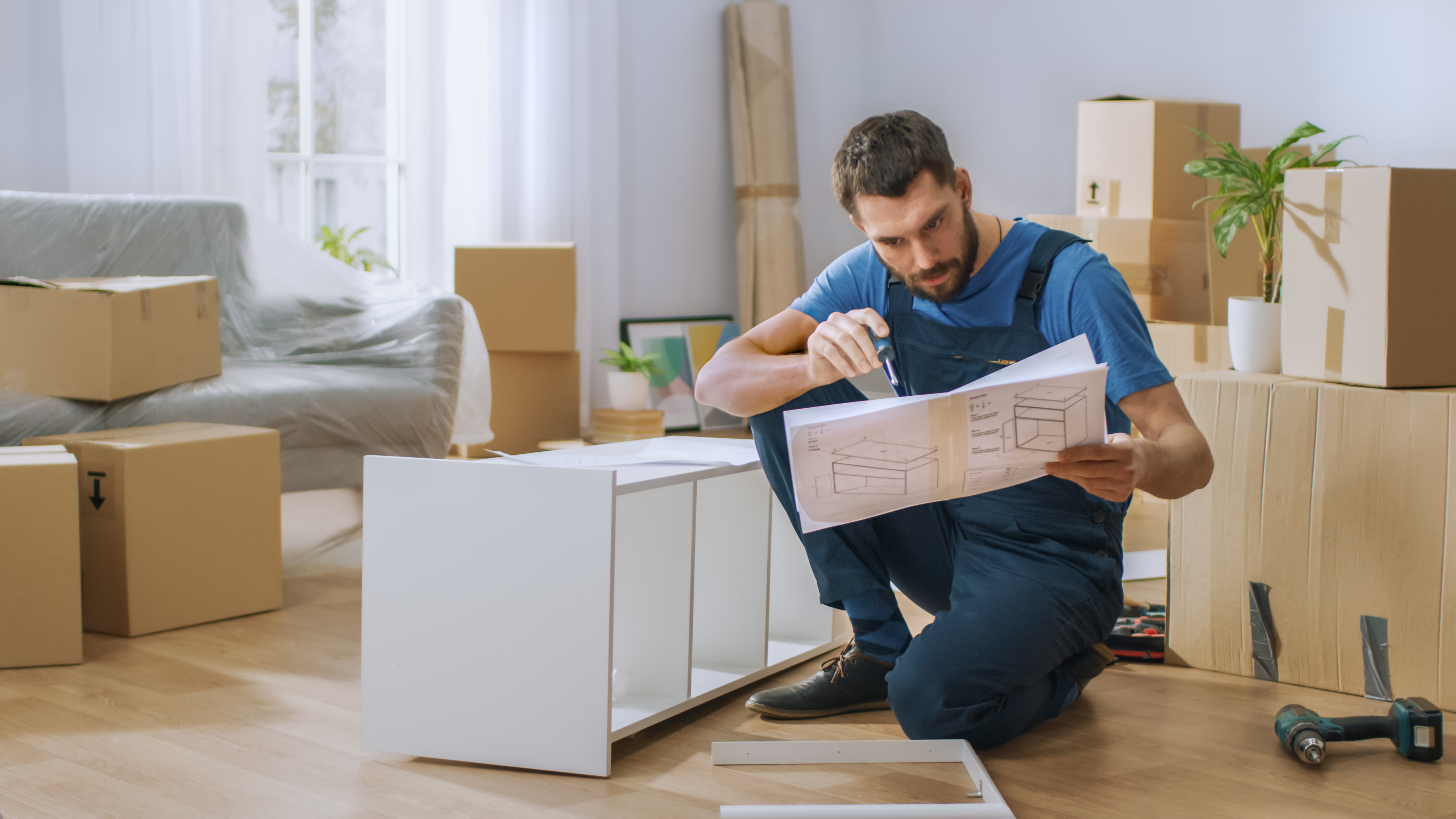 Man in blue overalls assembling white furniture in a room with cardboard boxes and plants around