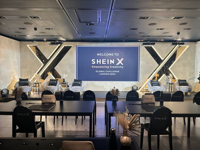 A conference room set up for the Shein X Global Challenge 2024 in London, featuring a stage with chairs, a backdrop sign, and branded tables