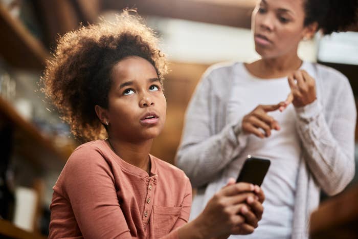 A frustrated young girl with a smartphone and an older woman gesturing behind her, looking concerned