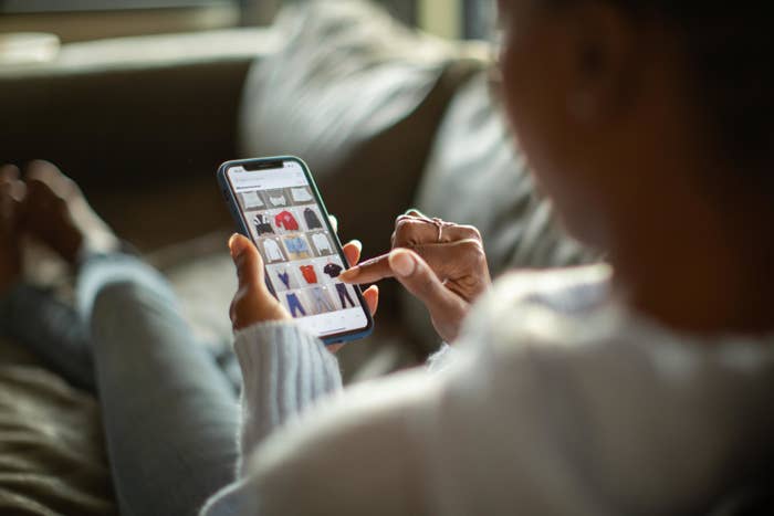 Person shopping online for clothes on a smartphone, browsing through sweaters and coats while sitting comfortably on a couch