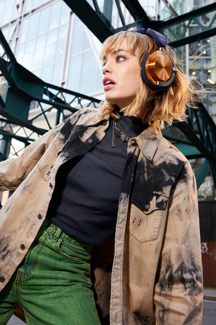 Seseorang mengenakan headphone besar, jaket denim tie-dye yang bergaya, turtleneck hitam, dan celana jins hijau. Struktur arsitektur modern terlihat di latar belakang
