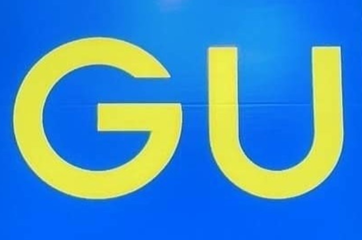 Large blue and yellow "GU" sign on a storefront with reflective surfaces and greenery in the background