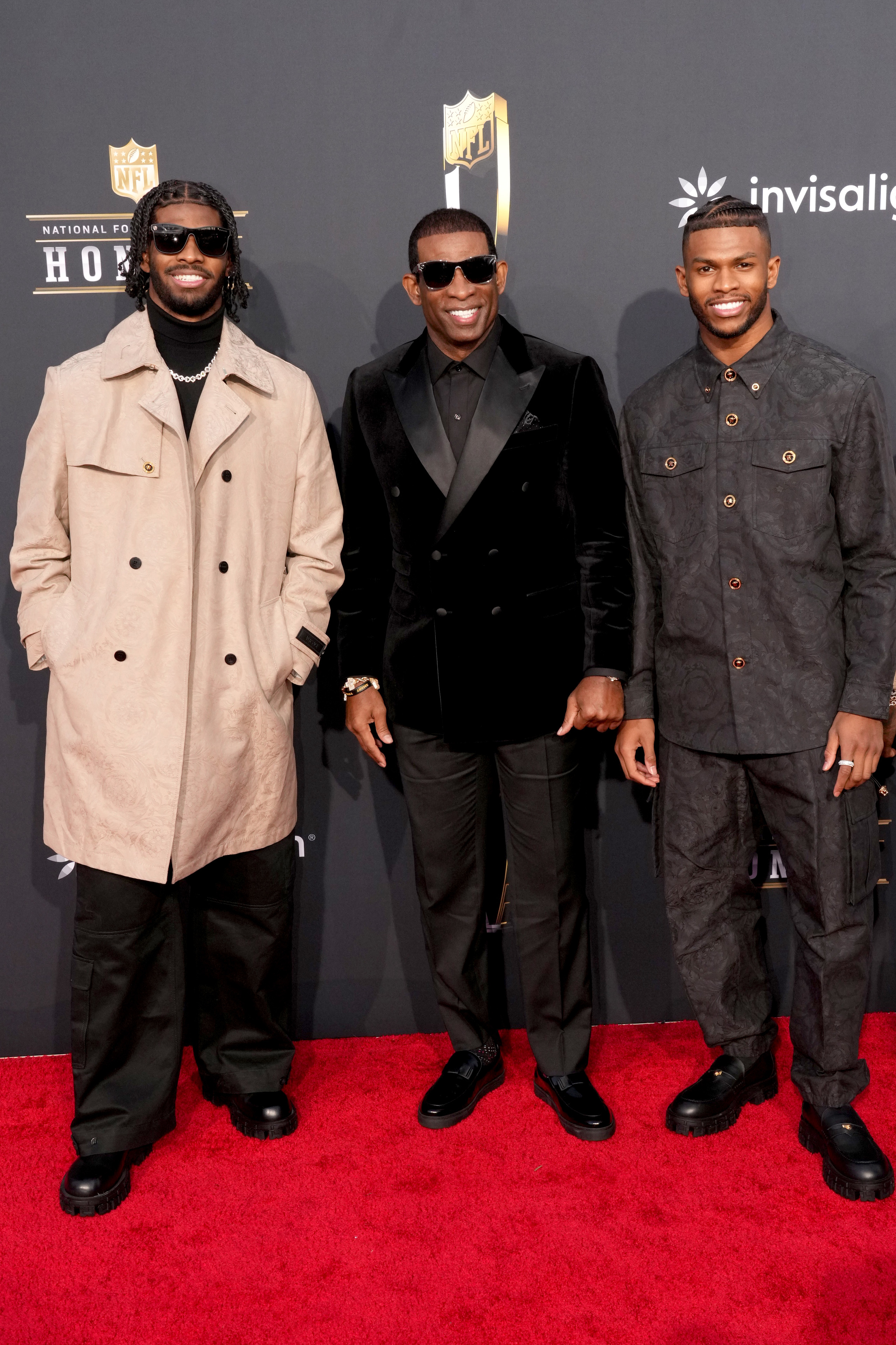 From left to right, Shedeur Sanders in a long coat, Deion Sanders in a black velvet suit, and Shilo Sanders in a patterned suit on the red carpet at an event