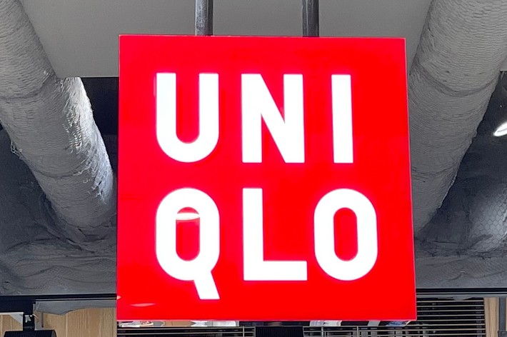 ユニクロの店内に吊るされた赤い看板には「UNIQLO」と書かれている。周囲には商品の棚が見える。