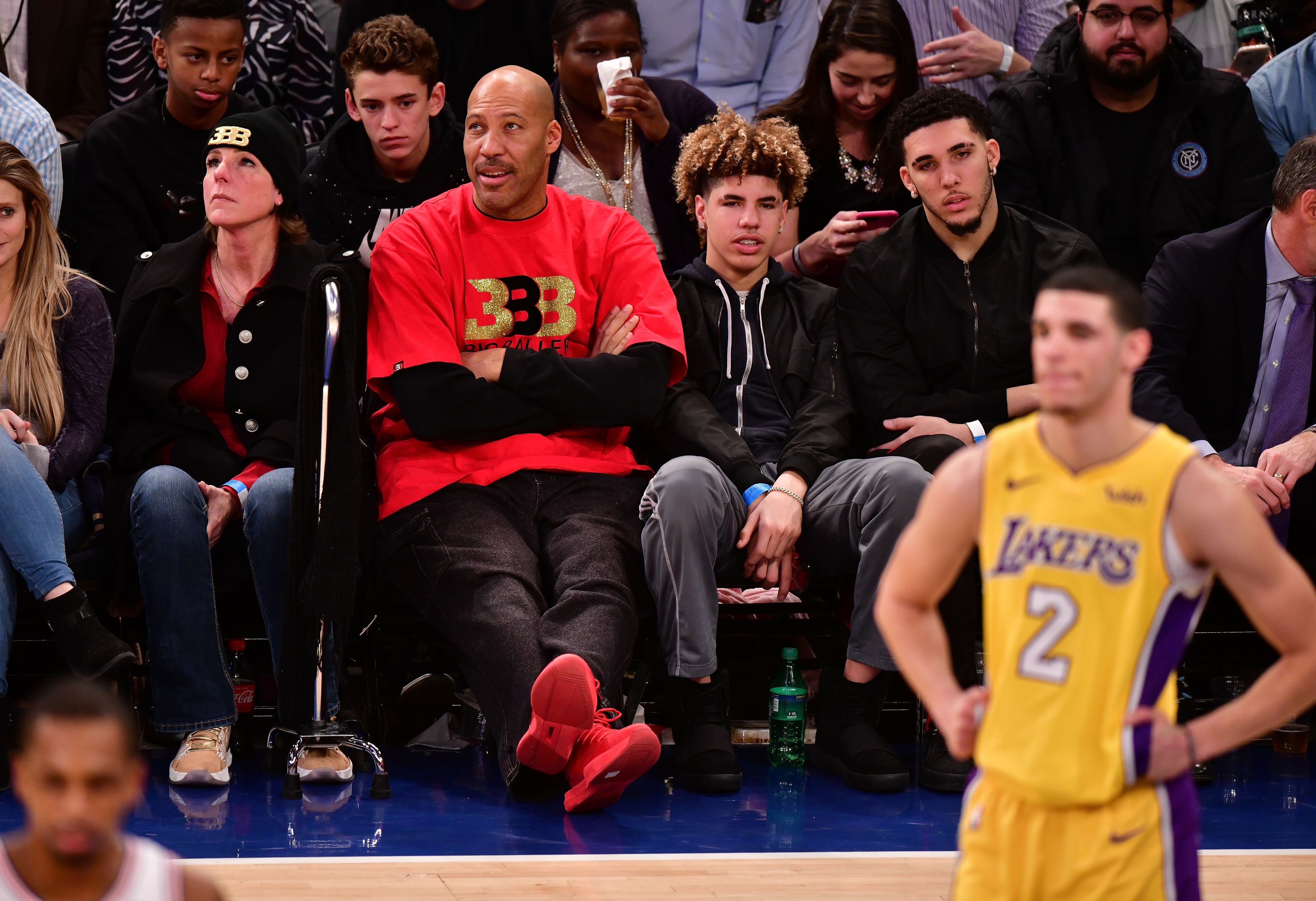 LaVar Ball in a red T-shirt sits courtside next to his sons LaMelo Ball and LiAngelo Ball. Lonzo Ball, in a Lakers jersey, is on the court