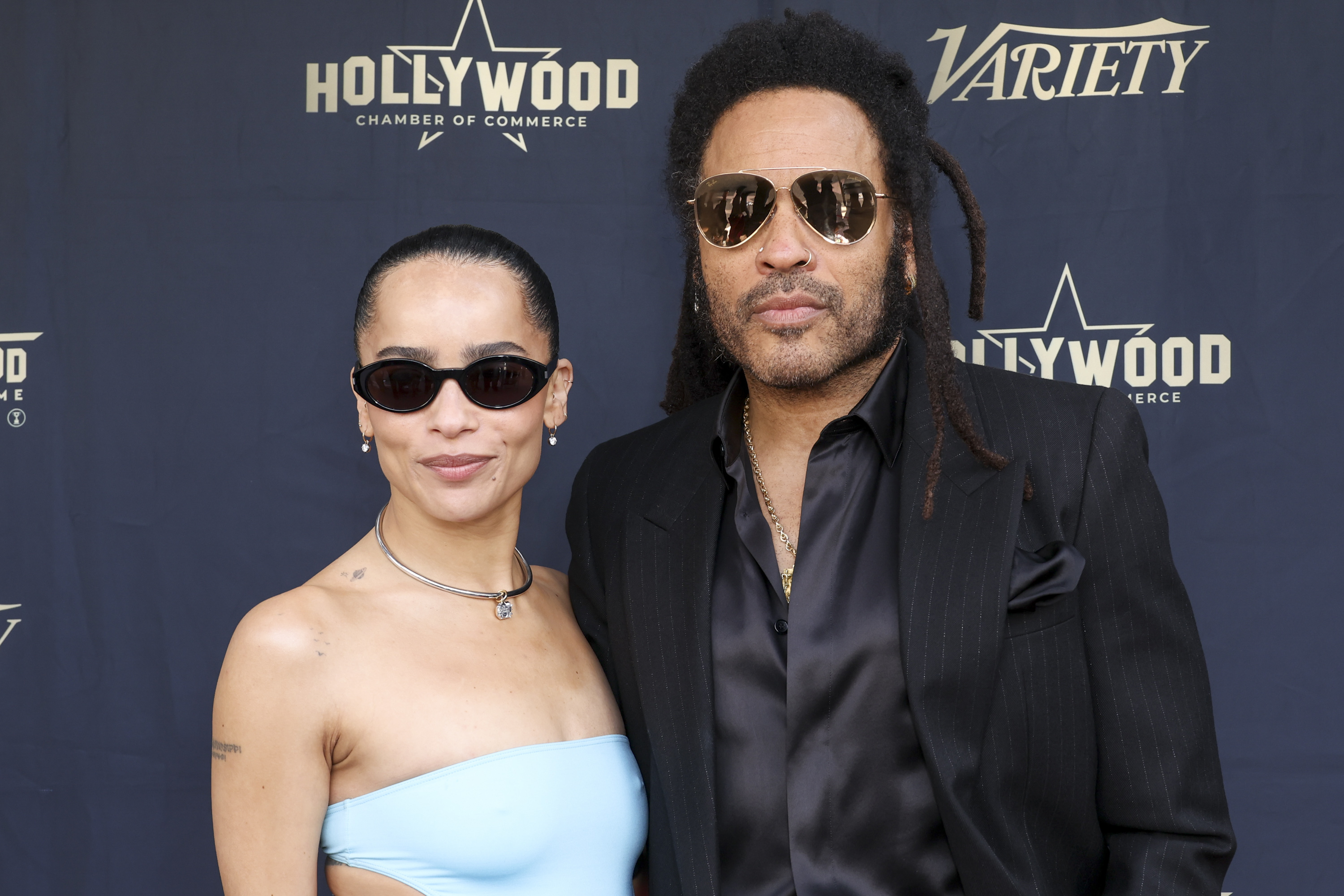 Zoe Kravitz and Lenny Kravitz stand in front of a Hollywood Chamber of Commerce backdrop. Zoe wears a sleek, strapless outfit and sunglasses; Lenny wears a dark suit and sunglasses