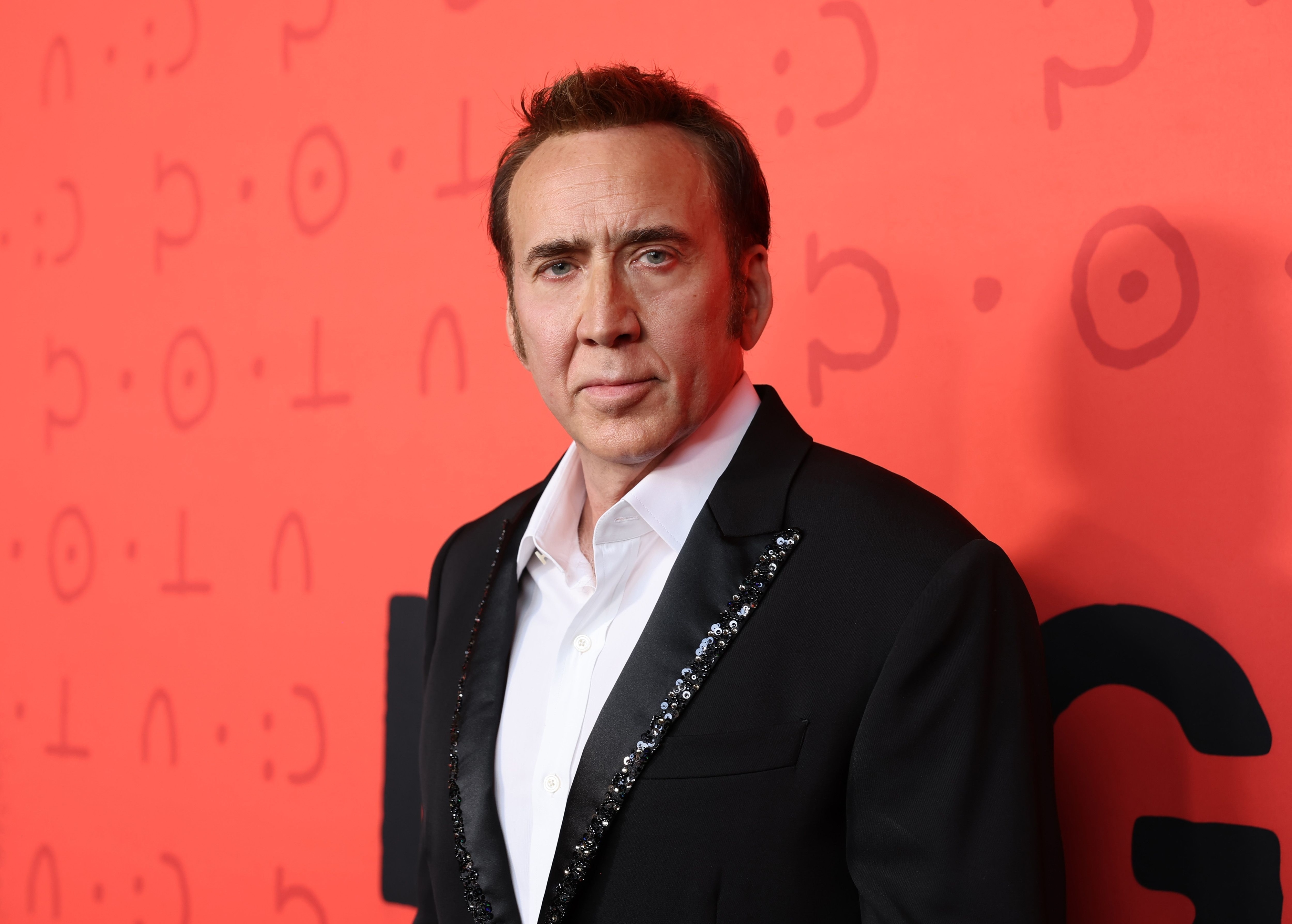 Nicolas Cage standing on a red carpet, wearing a black suit jacket with a white shirt, and embellished lapels