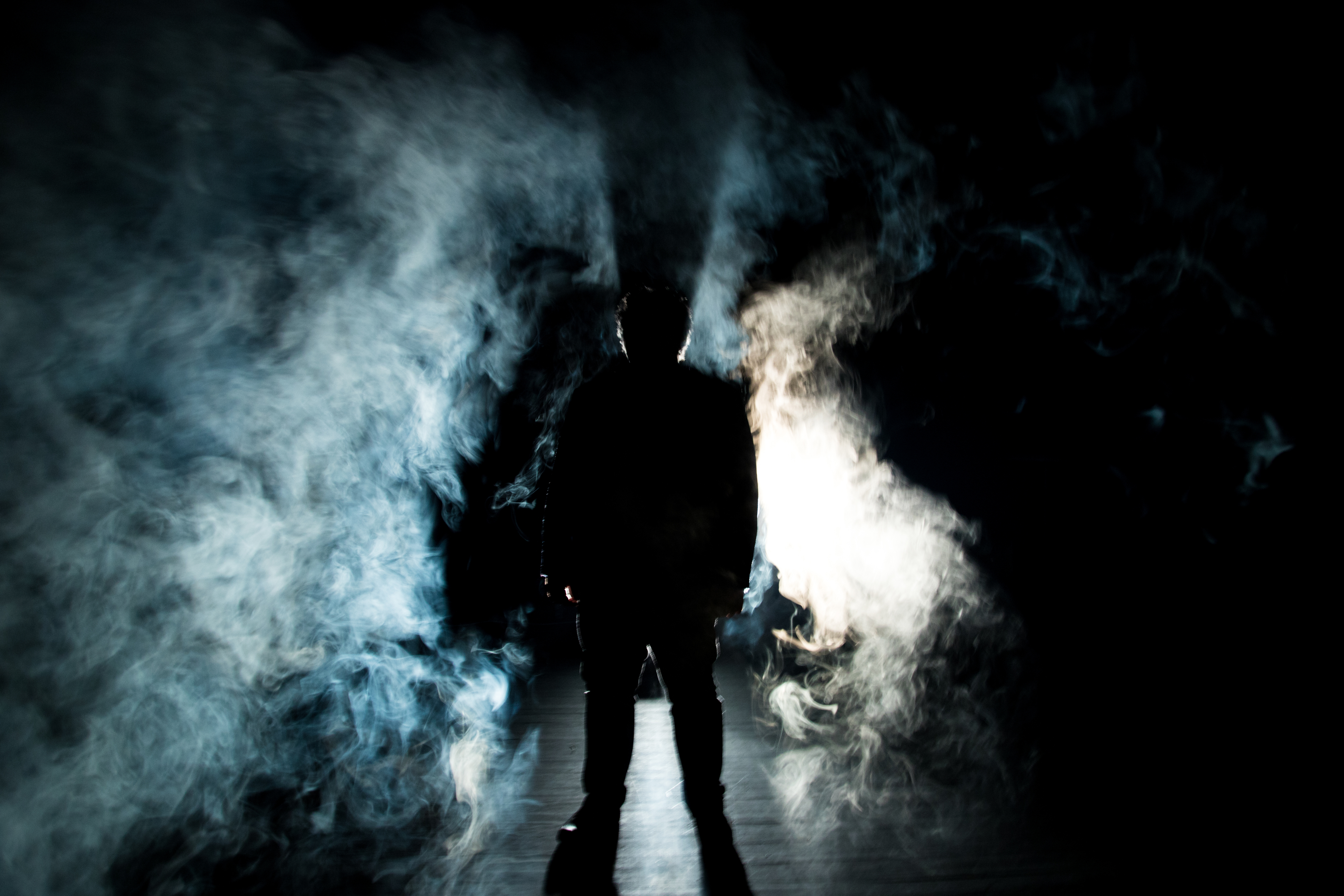 A person stands in a dramatic cloud of smoke with an intense light behind them, casting a silhouette. The person's identity is not visible