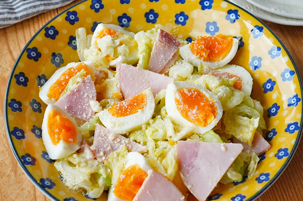 色とりどりの鮮やかな器に盛り付けられたハムとゆで卵のサラダです。背景にストライプの布が見えます。