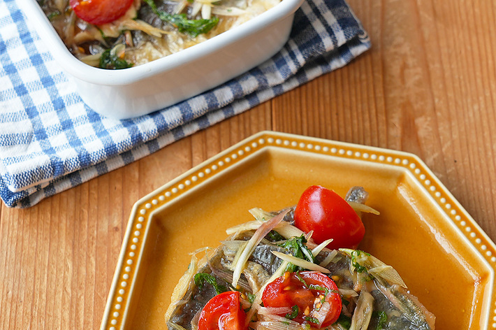 皿の上にトマトとハーブで飾られた魚料理が載っている。背景にはチェックの布と白い皿が見える。