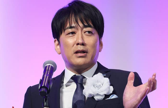 黒いスーツと青いネクタイを着た男性がトロフィーを持ちながらマイクの前で話している。