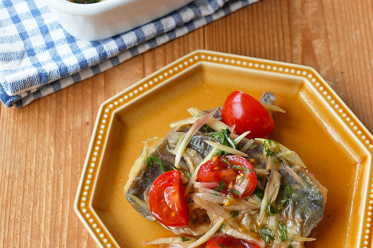 お皿に盛られた焼き魚と野菜の料理。隣には青いチェック柄の布の上に載った白い器が置かれている。