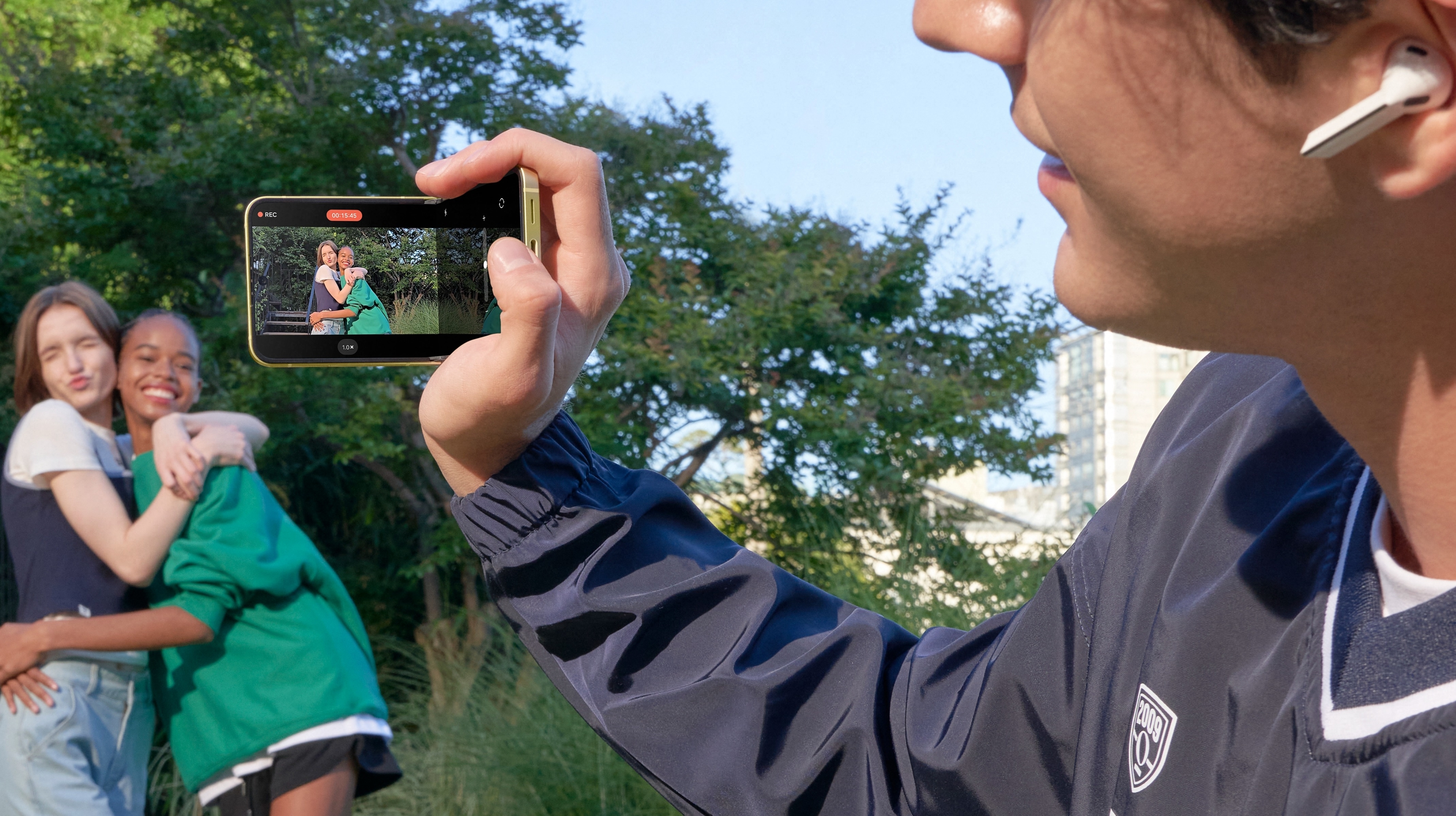A person wearing earphones and holding a smartphone takes a photo of two friends posing and hugging in a park