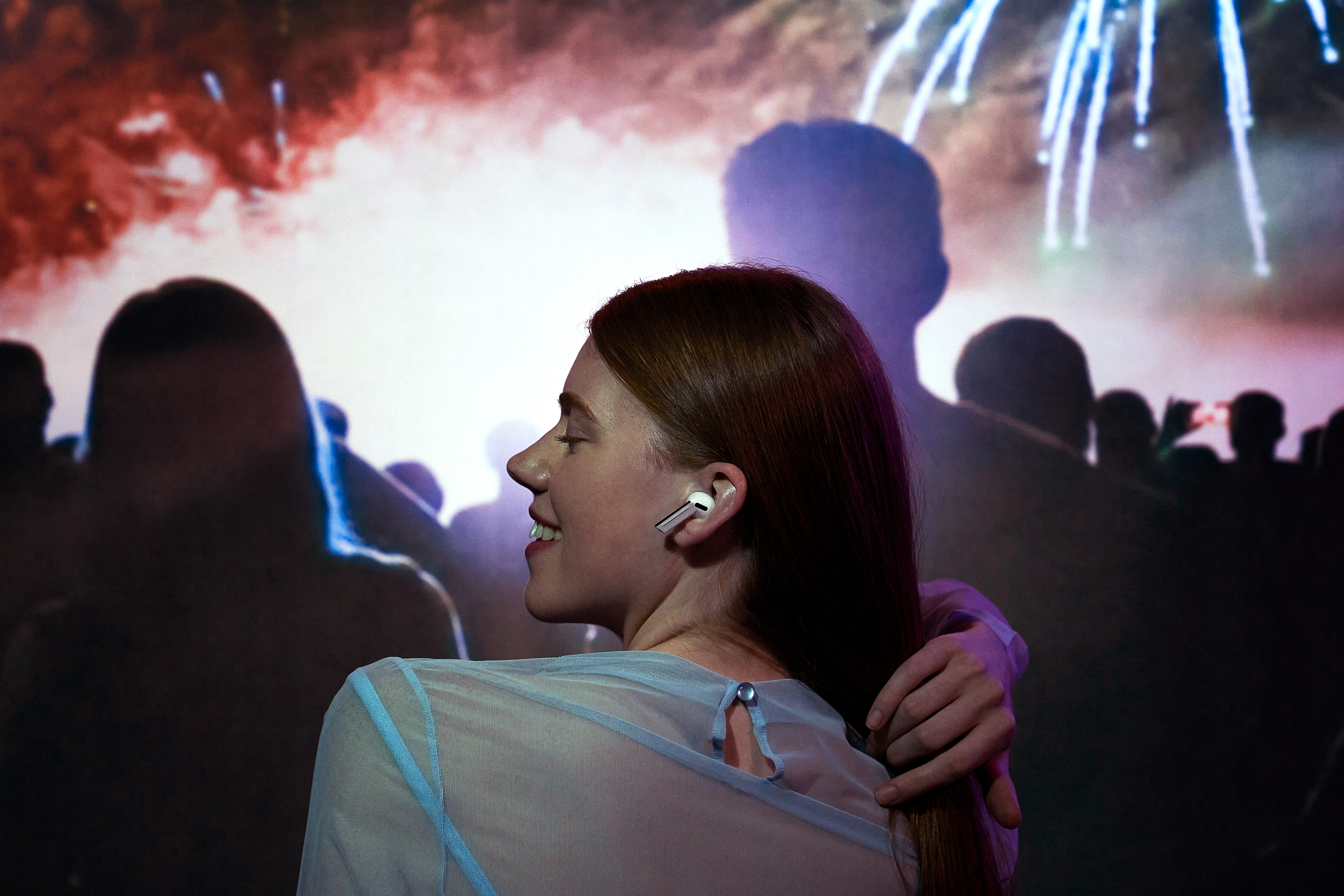 A woman with red hair smiles while wearing wireless earbuds in a crowd, with fireworks in the background