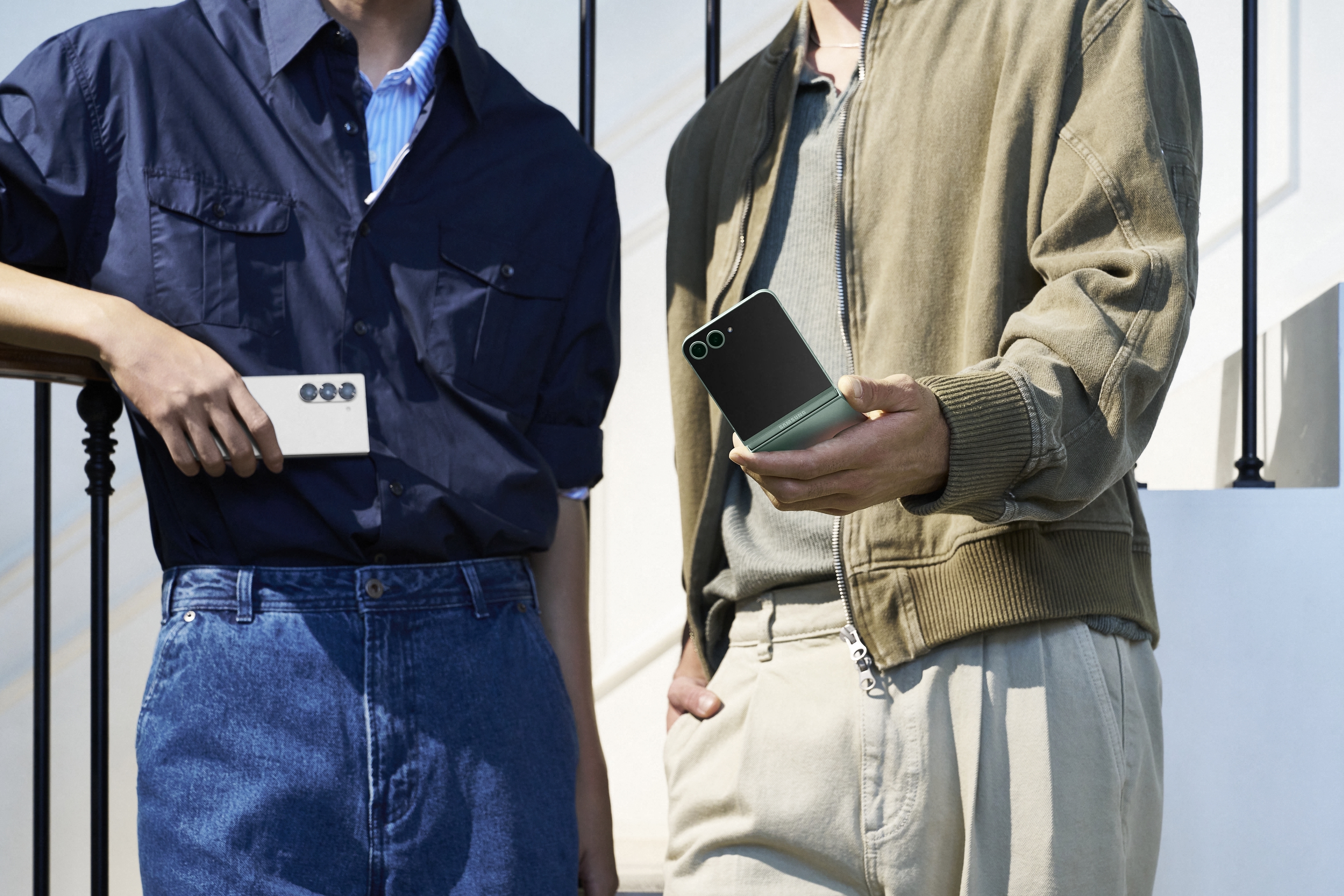 Two people holding Samsung Galaxy Z Fold 5 phones, one in green and one in silver, standing casually with stylish, relaxed clothing. Faces are not visible