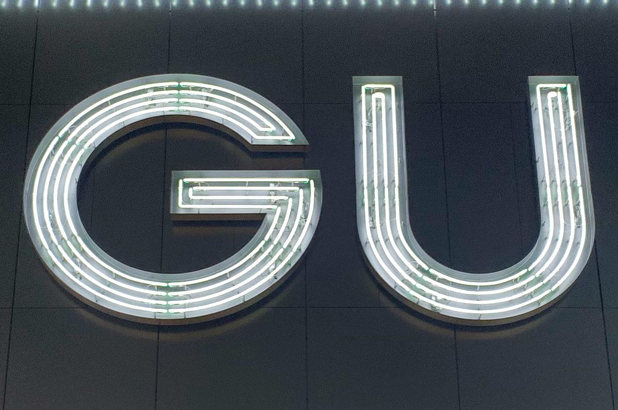 Illuminated "GU" sign on a building facade, composed of bright, evenly spaced horizontal lines