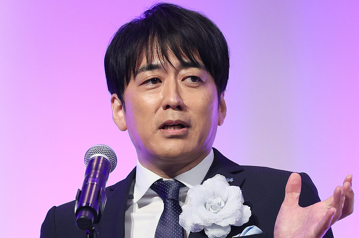 黒いスーツと青いネクタイを着た男性がトロフィーを持ちながらマイクの前で話している。