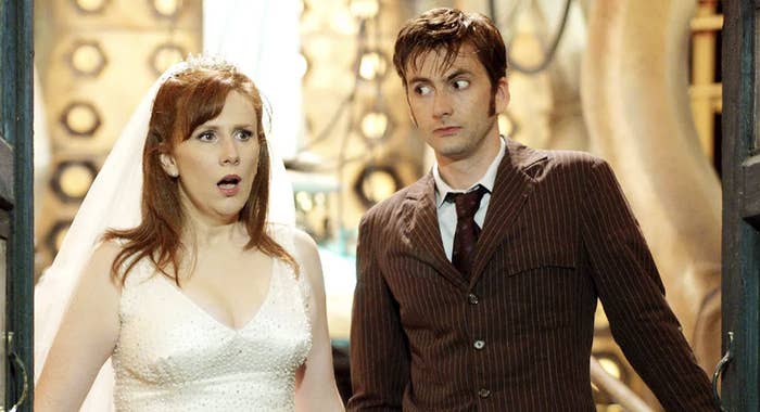 Catherine Tate in a wedding dress stands next to David Tennant in a pinstripe suit in a scene from Doctor Who, looking surprised