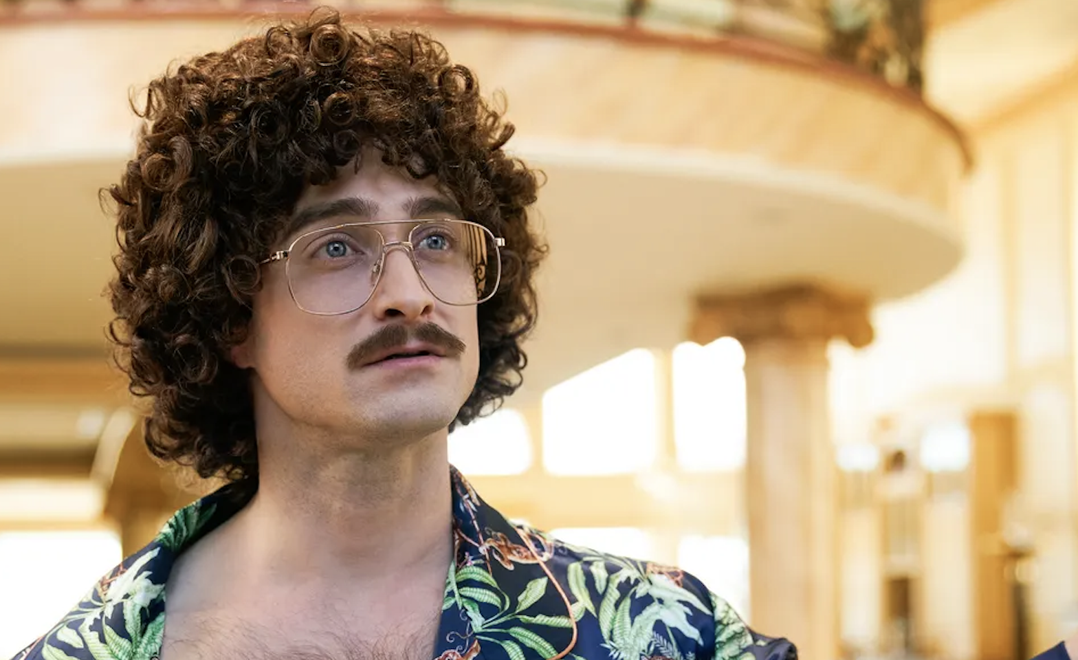 Daniel Radcliffe with curly hair, glasses, and a mustache, wearing a floral-patterned shirt
