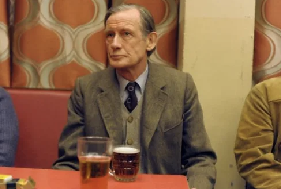 An older man sits at a table in a public setting, looking away from the camera with a thoughtful expression. A pint of beer is on the table in front of him