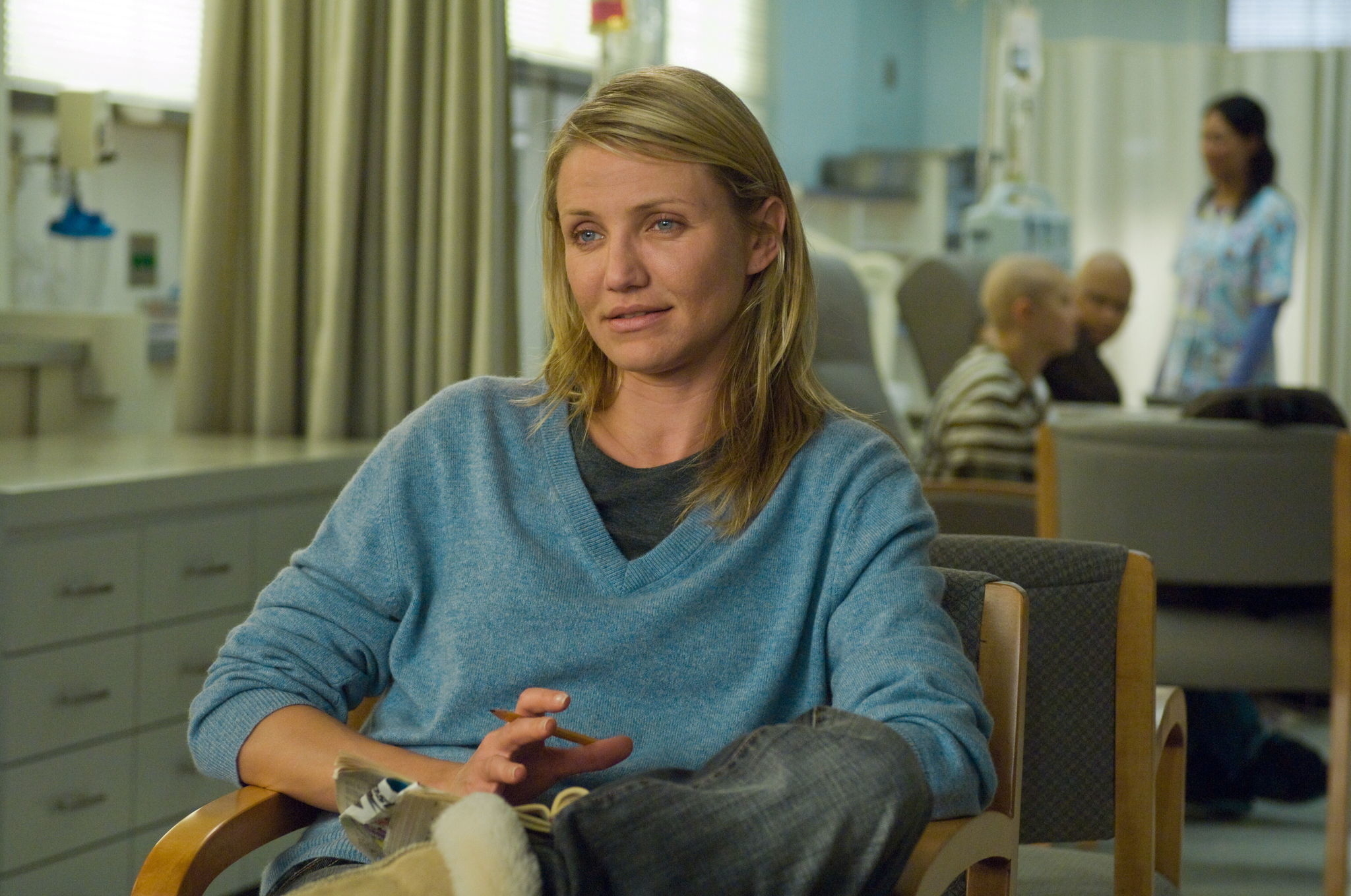 Cameron Diaz sits in a hospital room, wearing a casual sweater. In the background, a woman in a lab coat and patients, including children, are visible