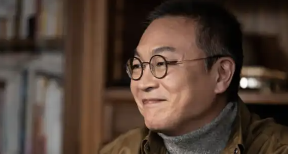Person smiling, wearing round glasses and a turtleneck sweater under a jacket
