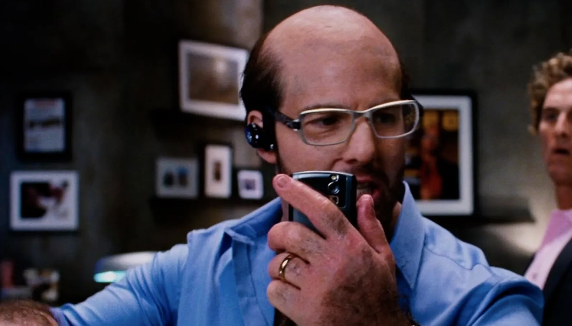 A man wearing glasses, holding a phone, speaks intensely into a walkie-talkie. Another person is partially visible in the background