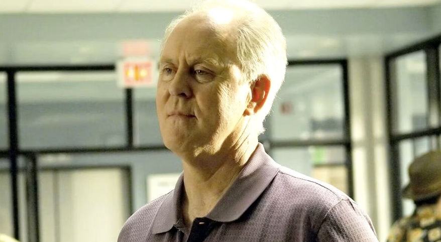 A man with short, light hair, wearing a collared shirt, is standing indoors in a hallway. The background includes windows and a wall with signage