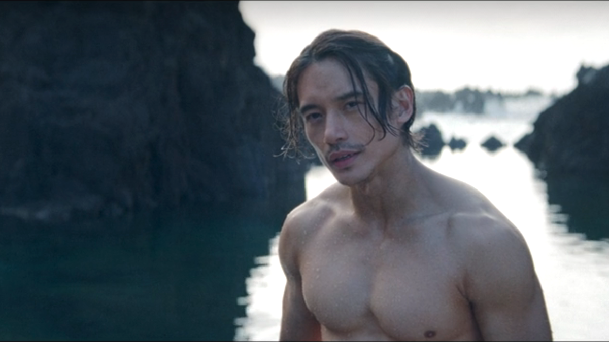 Mike Angelo, shirtless, stands in a serene outdoor setting with rocky cliffs and water in the background