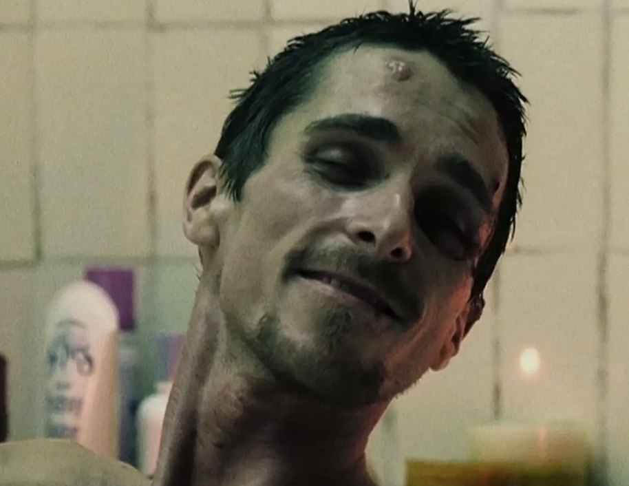 A person with dark hair smirking slightly, showing a gaunt physique and a few blemishes on the face, seen in a close-up shot with bathroom items in the background