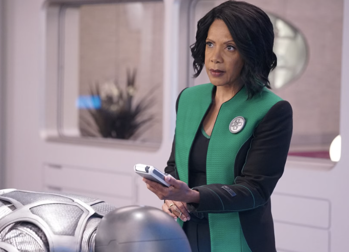 Penny Johnson Jerald in futuristic setting, wearing a green and black jacket, holding a device and appearing focused