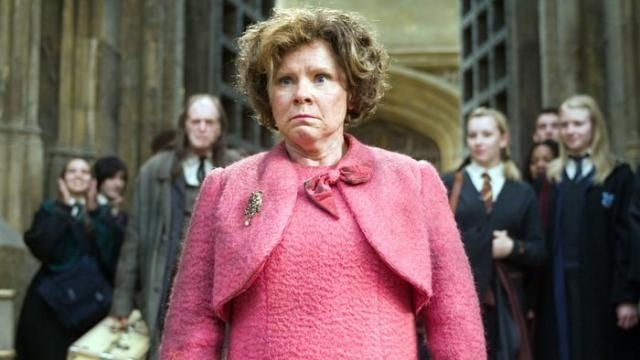 Imelda Staunton as Professor Umbridge stands sternly in a pink outfit in a scene from Harry Potter, with students and faculty visible in the background