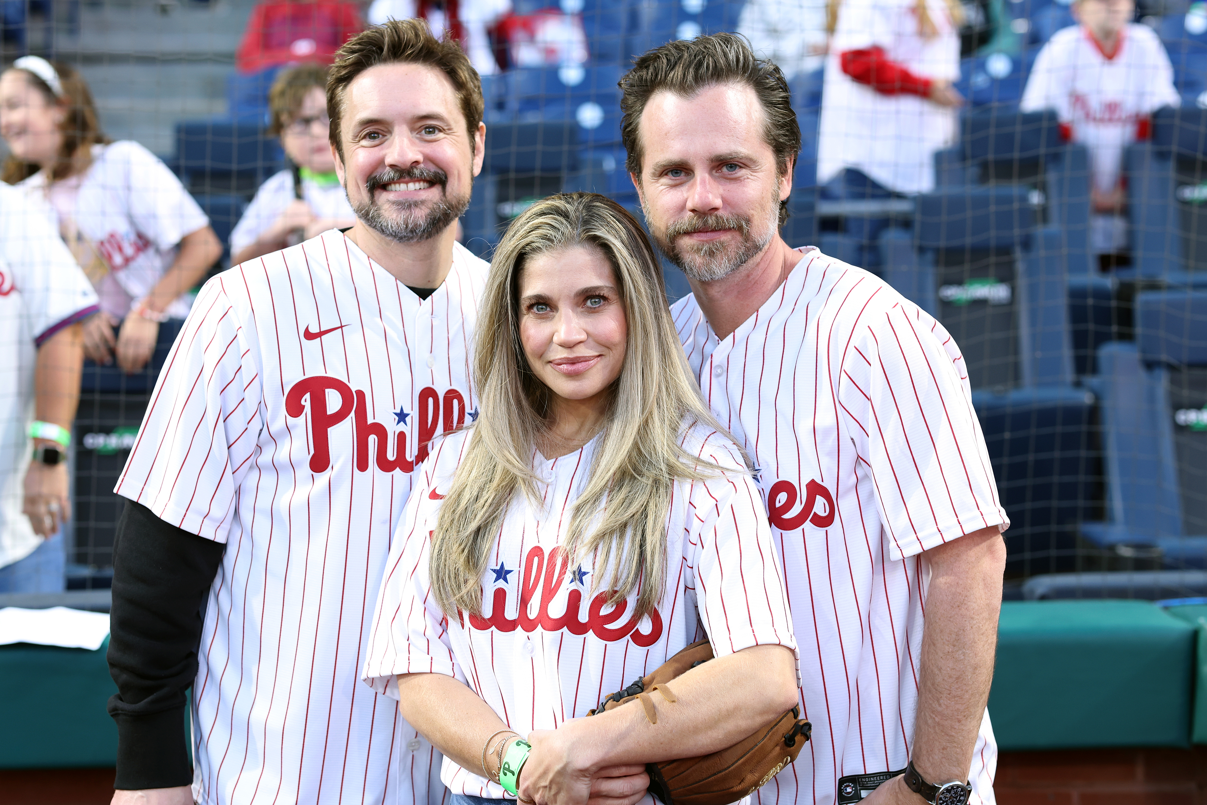 Rider Strong, Danielle Fishel, and Will Friedle wearing Phillies jerseys and standing together at a baseball stadium