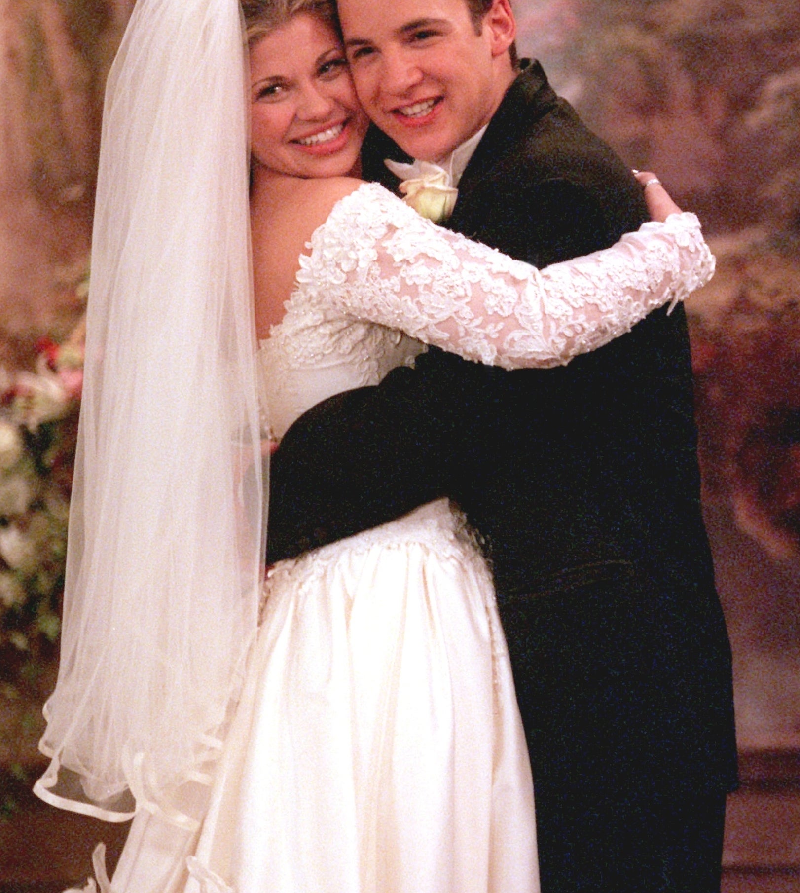 Danielle Fishel and Ben Savage in wedding attire, smiling and embracing, with a floral backdrop