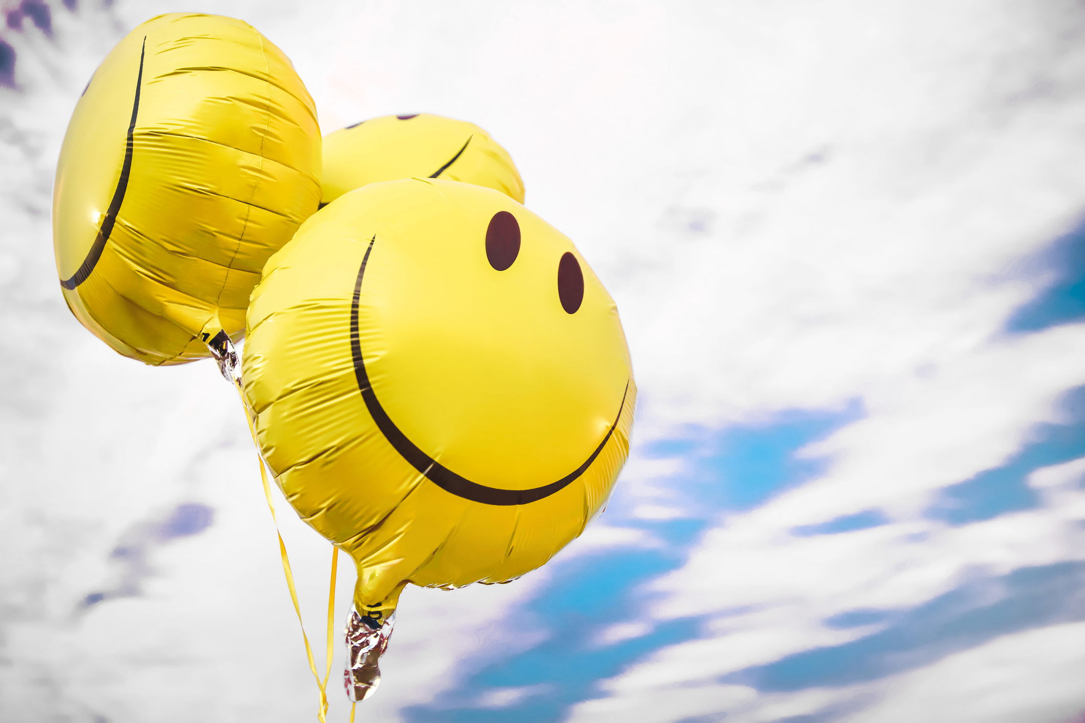 Three yellow balloons with smiley faces float against a partly cloudy sky