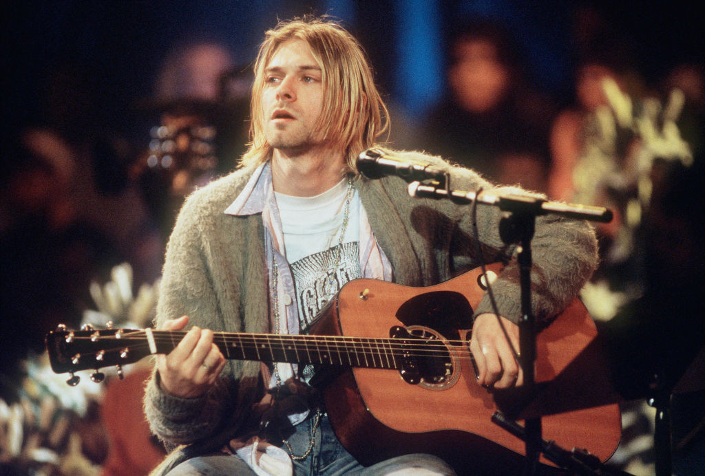 Kurt Cobain sits with a guitar during an MTV Unplugged performance. He is wearing a casual outfit including a cardigan and a t-shirt