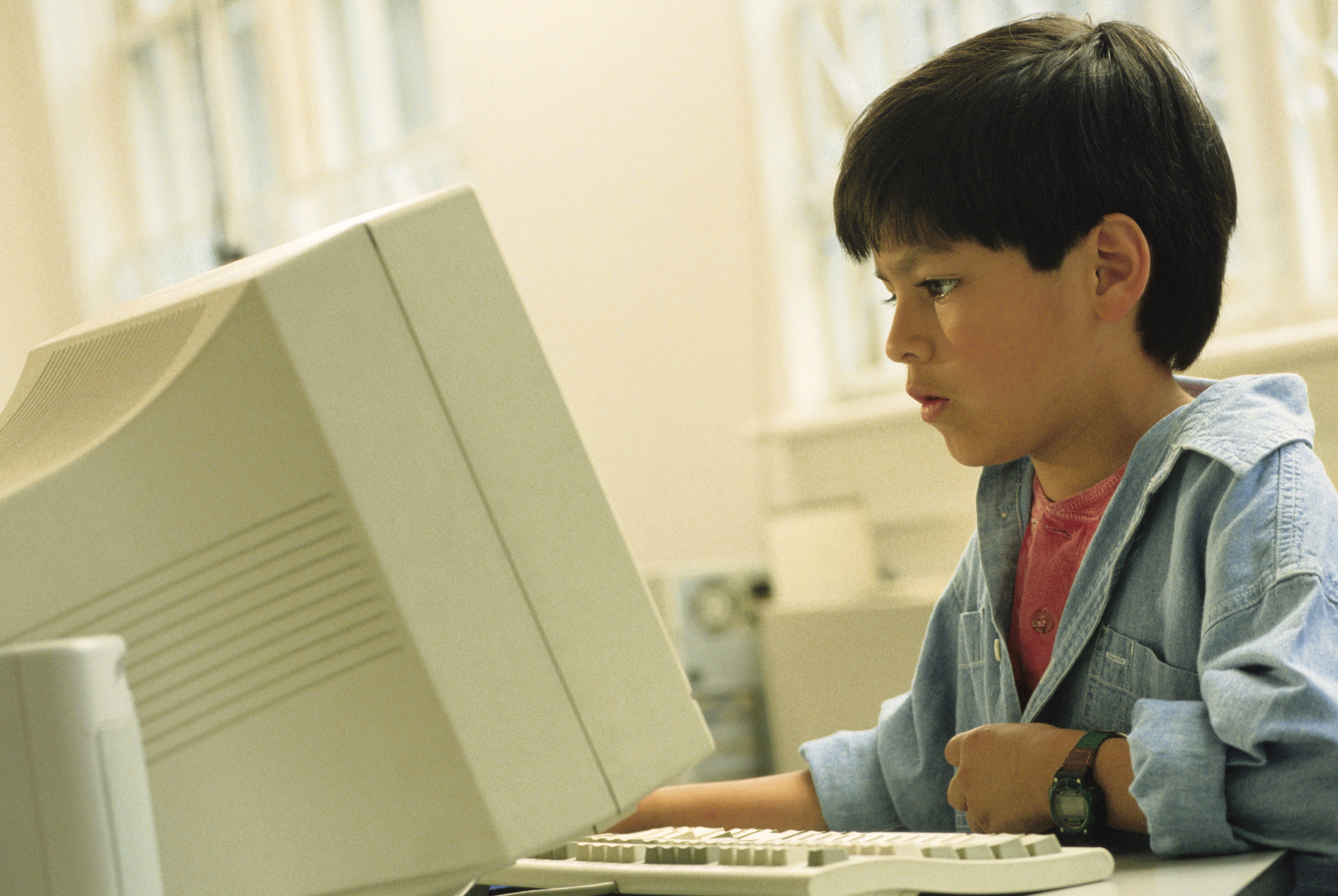 A young boy is seated at a desk, focused on a large, old-style computer monitor, suggesting a scene from the 1990s technology era