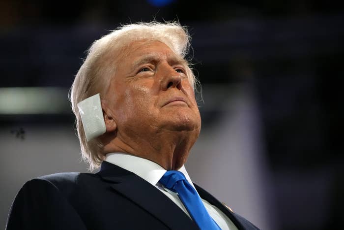 Donald Trump looking upwards with a stern expression, wearing a dark suit and blue tie, with a small bandage on the side of his head