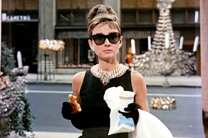 Audrey Hepburn in a classic black dress, pearls, gloves, sunglasses, holding a pastry in one hand and a coffee cup in the other, from "Breakfast at Tiffany's."