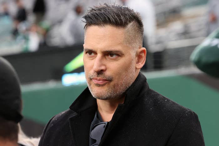 Joe Manganiello with short hair and a beard, wearing a coat and sunglasses around his neck, at an outdoor event