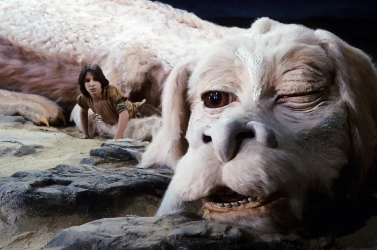 A boy (Atreyu) rests beside a large, white, furry dragon (Falkor) on a rocky surface from a scene in "The NeverEnding Story."