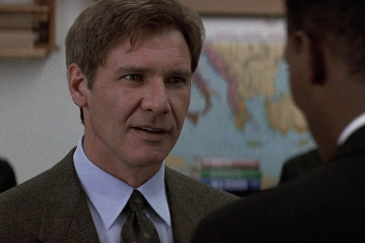 Harrison Ford in a suit and tie is speaking to another person who is out of focus. A wall map and shelves are in the background