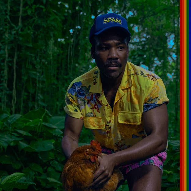 Donald Glover wearing a floral shirt and a SPAM cap, holding a chicken, stands in a lush, jungle-like setting